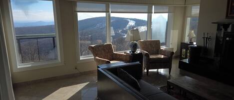A spacious living room with a large window offering a mountain view. There is a sofa, a chair, and a small table with a lamp.