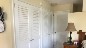 A hallway with white louvered doors, a wooden nightstand with a lamp, and a bed with a headboard.