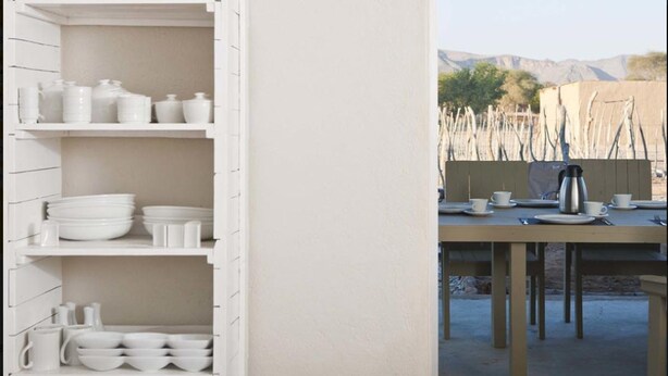 A dining area with a table set for two, a white cabinet with dishes, and a view of a mountainous landscape.