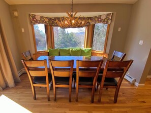 Ein Essbereich mit einem großen Holztisch und Stühlen, einer Chandelier und Blick auf Bäume durch große Fenster.