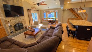 A spacious living room with a stone fireplace, a large brown sectional sofa, a wooden coffee table, and a dining area with a chandelier.