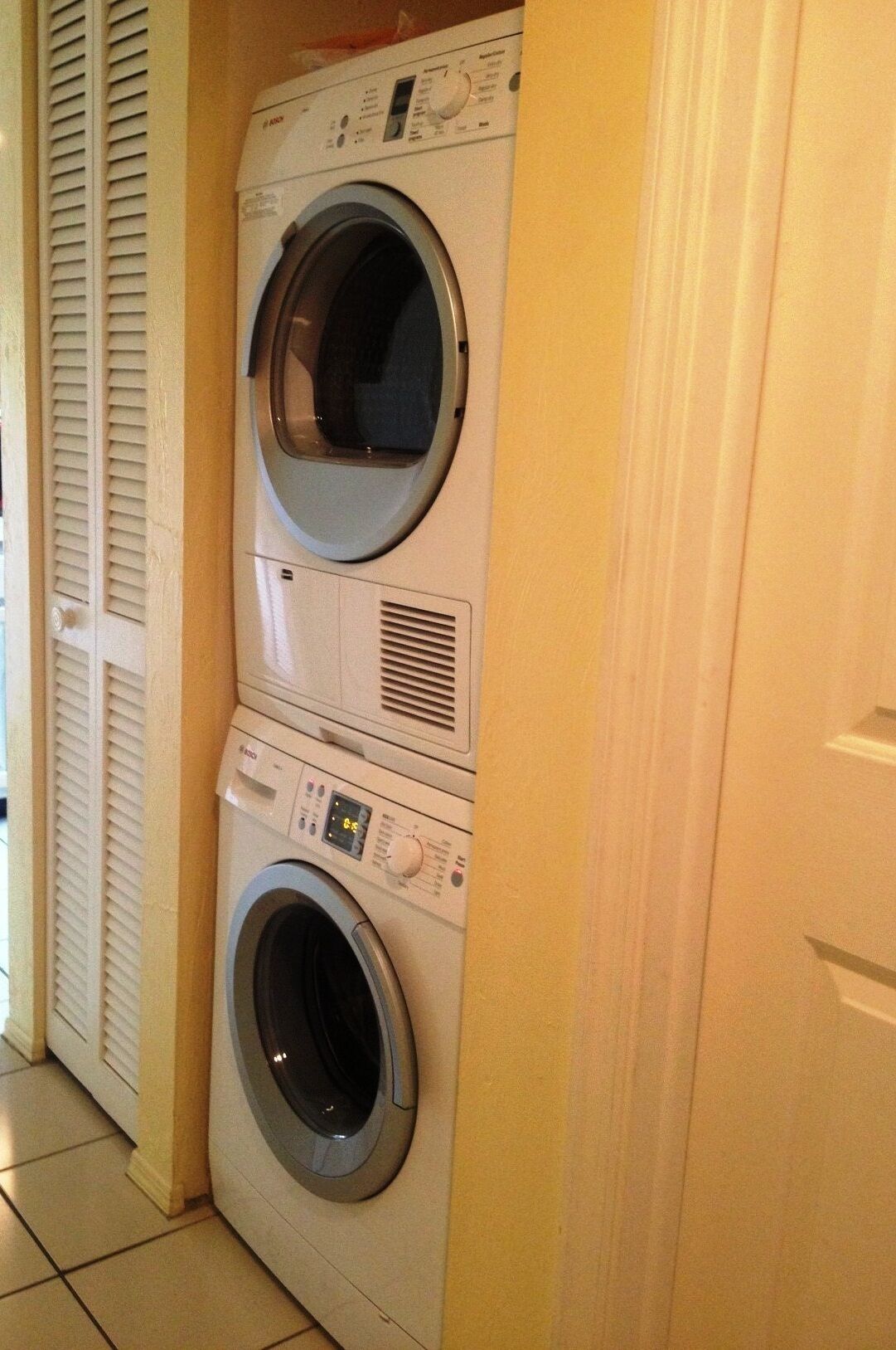 A front-loading washing machine and dryer stacked in a laundry room with a door and a window with blinds.