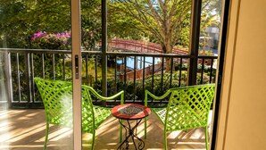 A balcony with two green chairs and a small table, overlooking a garden with a red bridge and trees. - Park Shore Resort #258 Top loc. Spectacular view, Beach, Tennis, Pickleball, Gym (Naples)