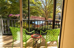 A balcony with two green chairs and a small table, overlooking a garden with a red bridge and trees. - Park Shore Resort #258 Top loc. Spectacular view, Beach, Tennis, Pickleball, Gym (Naples)