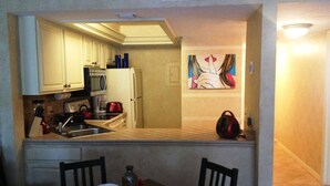 A compact kitchen with white cabinets, a microwave, and a refrigerator. There is a dining table with chairs and a painting on the wall. - Park Shore Resort #258 Top loc. Spectacular view, Beach, Tennis, Pickleball, Gym (Naples)