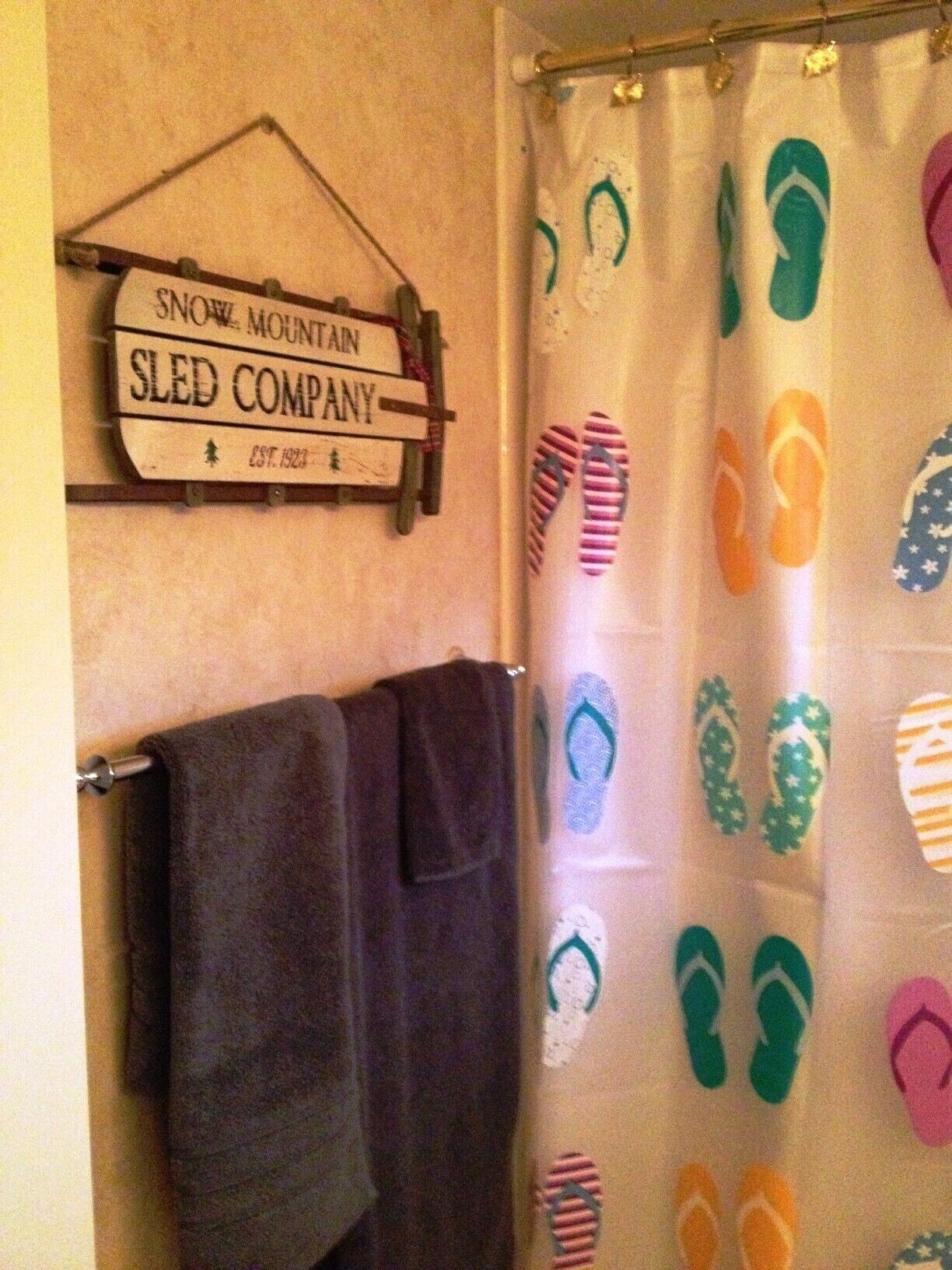 A bathroom with a shower curtain featuring a flip-flop pattern, a sign above the shower reading "SNOW MOUNTAIN SLED COMPANY EST. 1923", and a dark towel hanging on a rack.