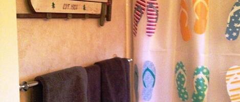 Un baño con cortina de ducha estampada con un patrón de chanclas, un cartel sobre la ducha que dice "SNOW MOUNTAIN SLED COMPANY EST. 1923" y una toalla oscura colgada en un perchero.