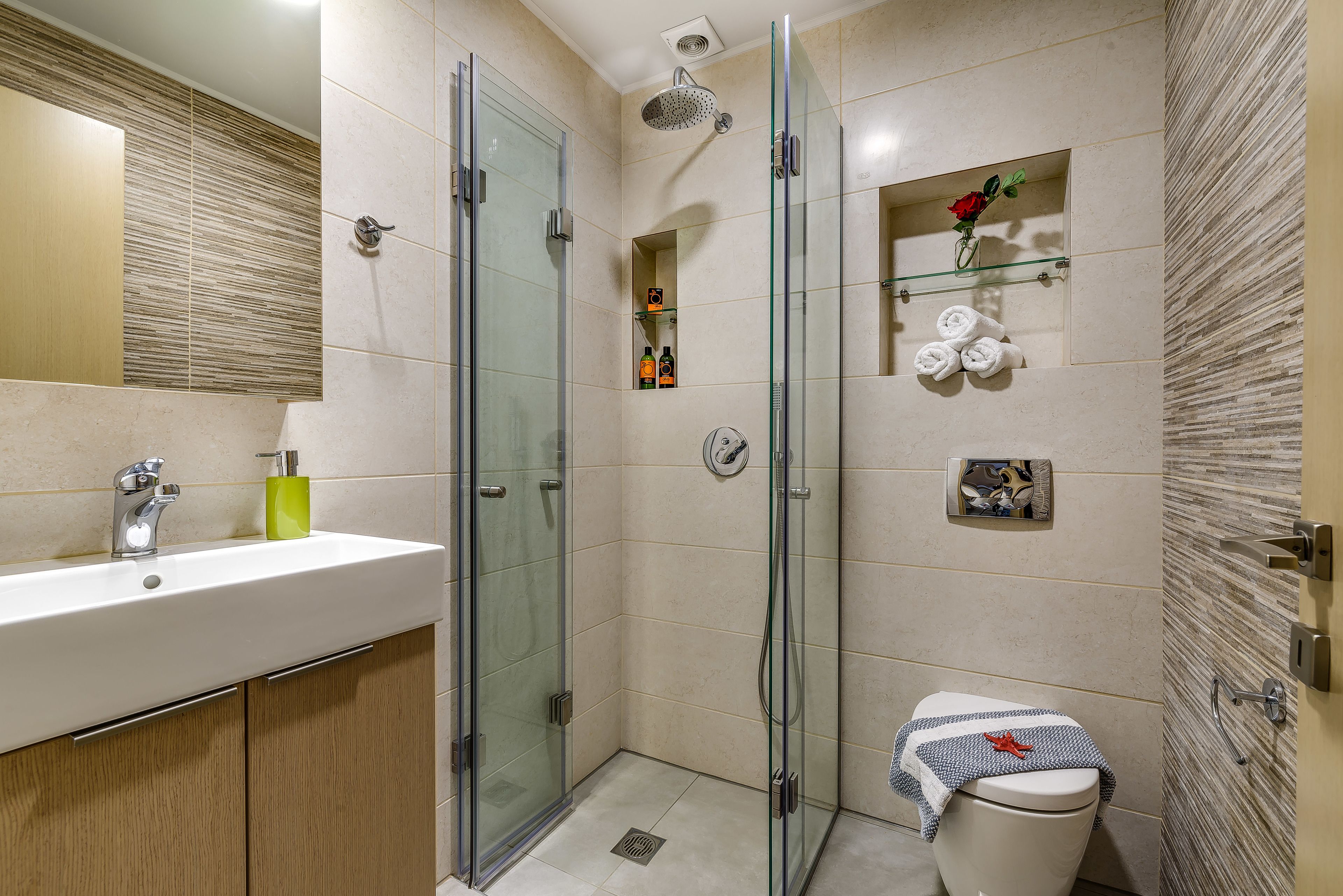 Un baño moderno con ducha de vidrio, lavamanos con espejo e inodoro con una toalla y un objeto decorativo.