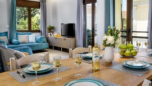 A modern living room with a sofa, dining table set with wine glasses and plates, a TV, and a balcony with a view of trees. - City Breeze Apartment (Chania)