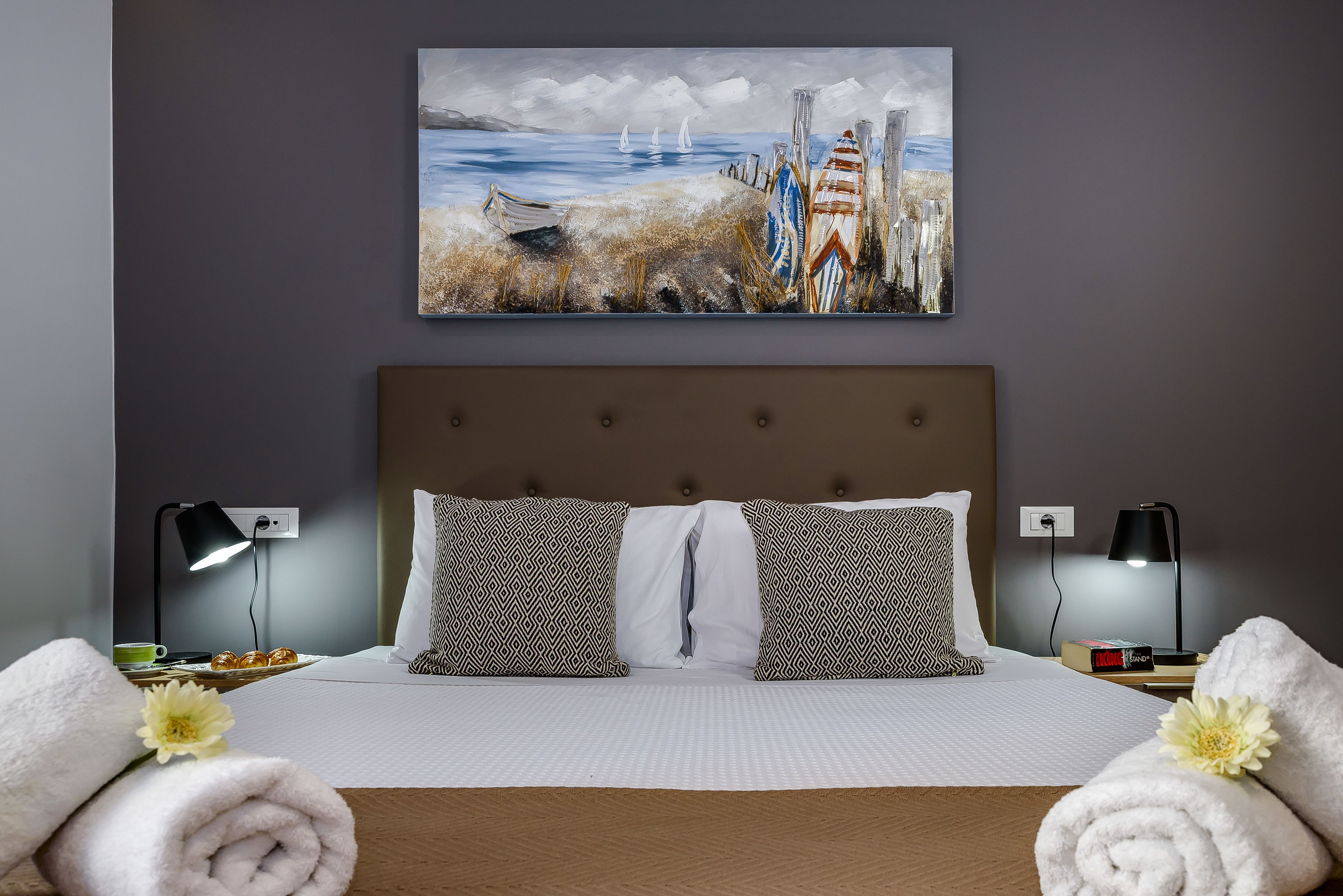 A neatly made bed with patterned pillows, a painting of a beach scene above it, and two bedside lamps.