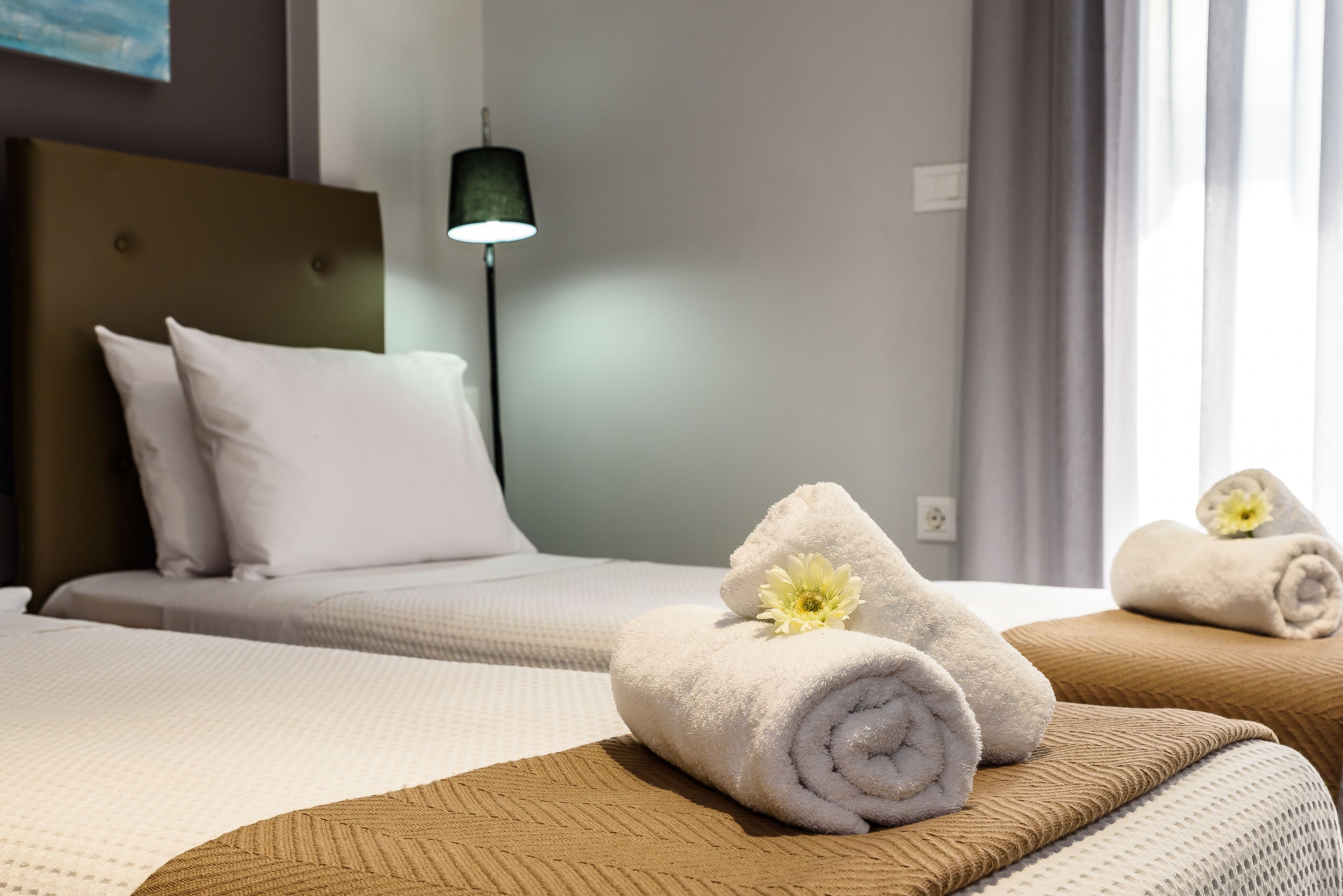 Camera d'albergo con due letti, ognuno con biancheria da letto bianca e una testiera. Ci sono degli asciugamani con dei fiori sui letti, e una lampada è visibile sul lato sinistro.