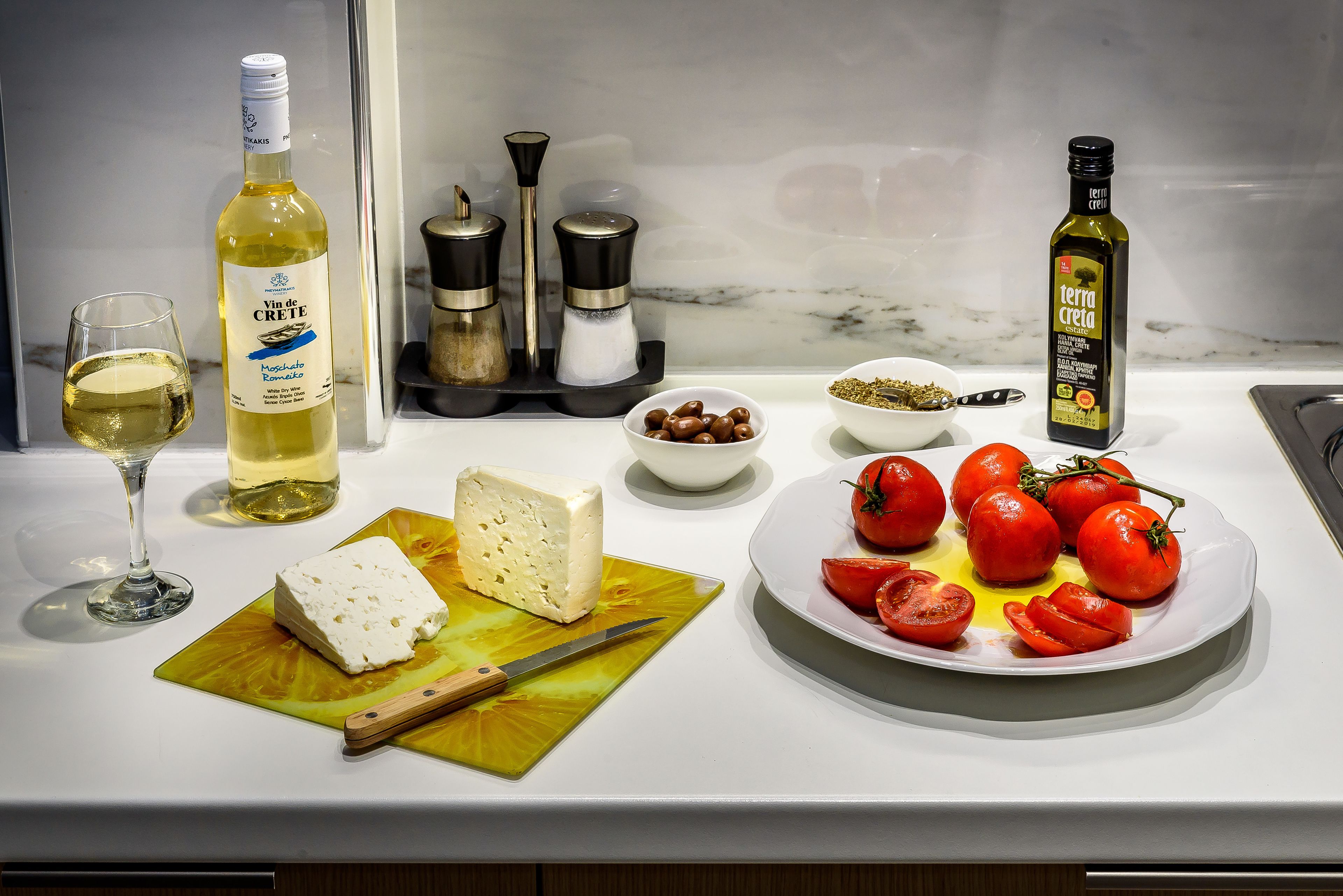 Un comptoir de cuisine avec une bouteille de vin blanc, un verre, un morceau de fromage, un couteau, une assiette de tomates et une bouteille d’huile d’olive.