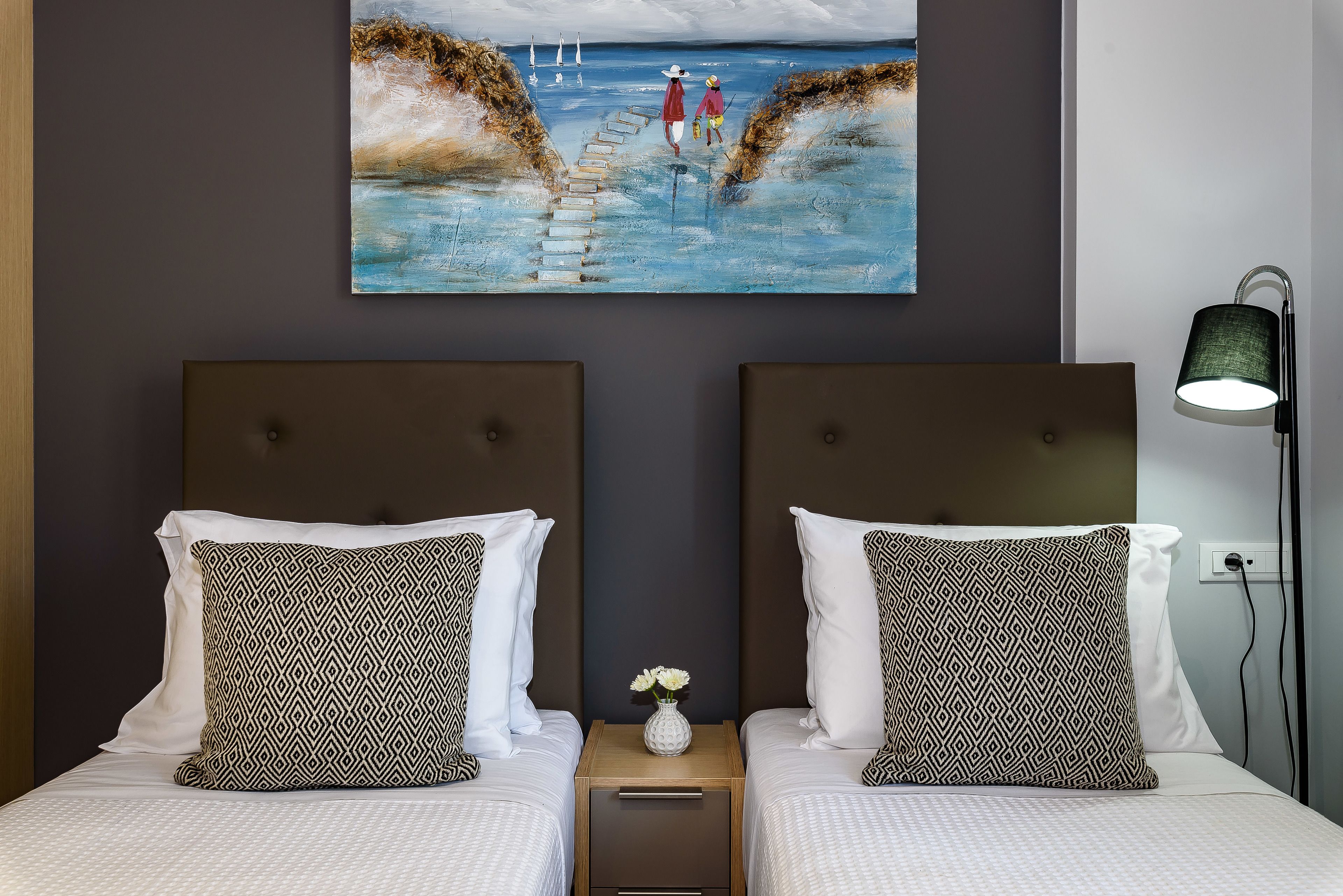A hotel room with two beds, a painting of a beach scene, a nightstand with a vase of flowers, and a lamp.