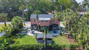 A two-story house with a patio area, surrounded by lush greenery and palm trees. - Folly Vacation 114B Townhouse, perfect family vacation, pets welcome! (Folly Beach)