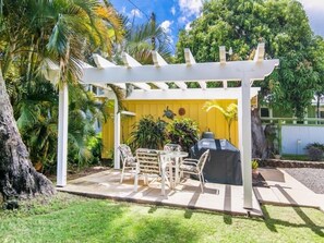 Um alpendre coberto ao ar livre com pérgola branca, churrasqueira e mesa com cadeiras.