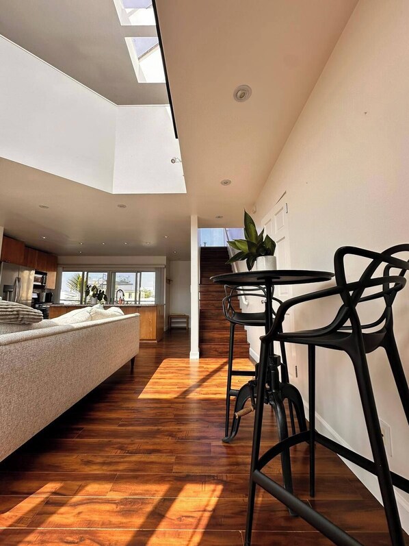 A spacious living area with a high ceiling, wooden flooring, a sofa, a dining table with chairs, and a staircase leading to an upper level.