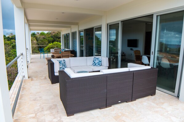 A modern outdoor living area with a large sofa and a dining table, connected to a glass sliding door leading to a balcony.
