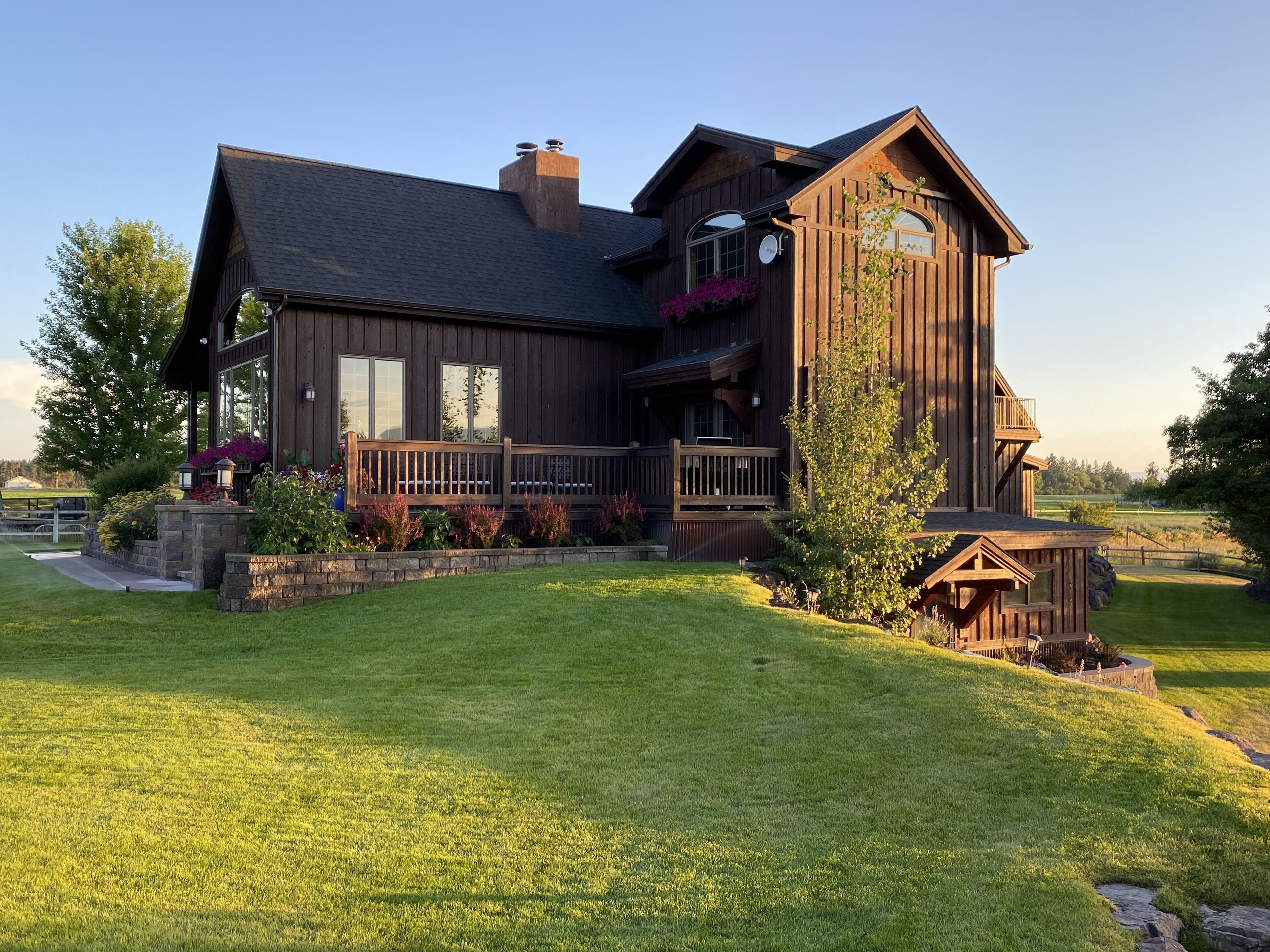 A large, two-story wooden house with a balcony, surrounded by a well-maintained lawn and flowering plants.