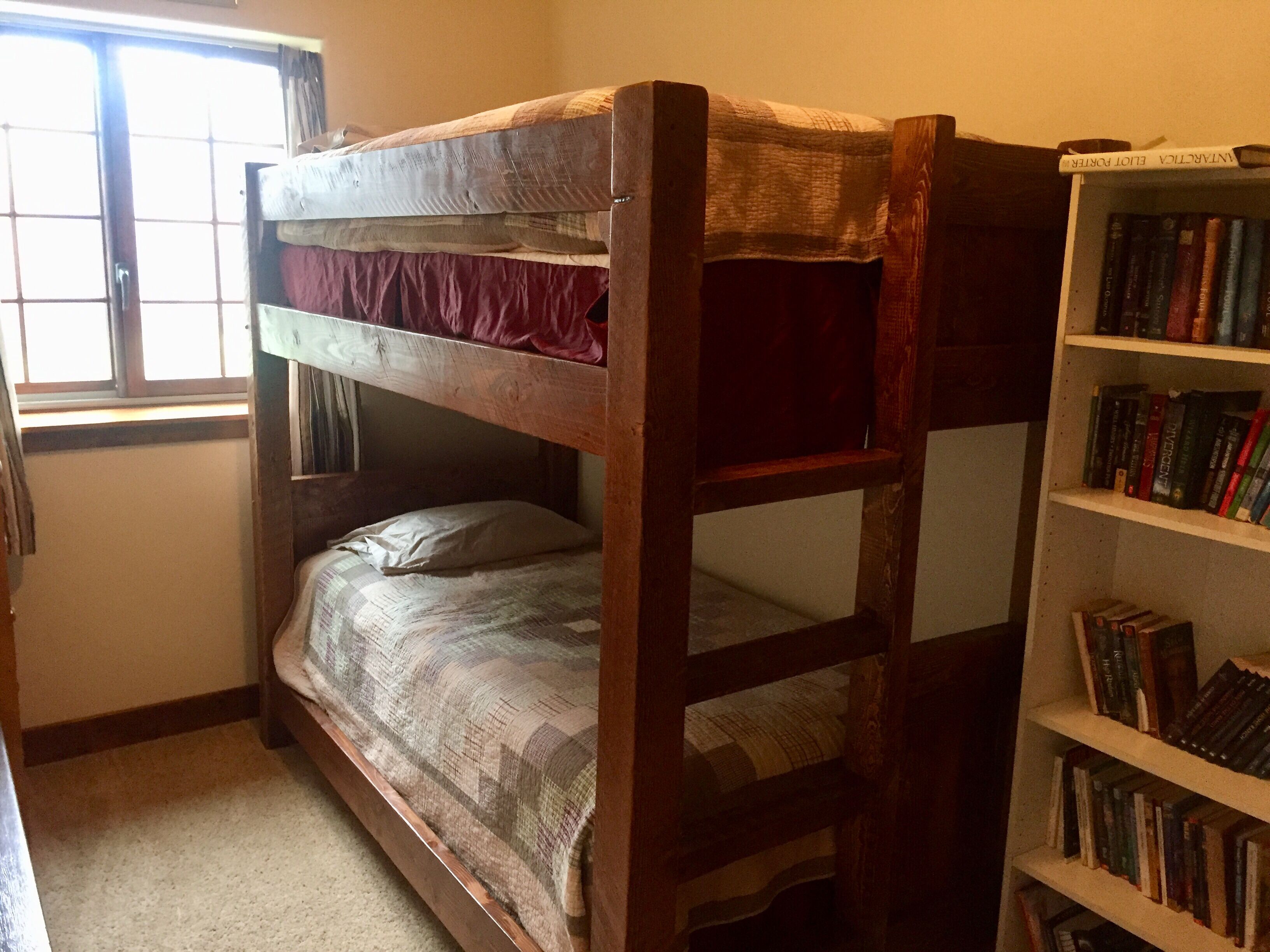 Un lit superposé avec un matelas rouge et une tête de lit en bois. Une bibliothèque remplie de divers livres se trouve à droite. Une fenêtre avec des rideaux est visible à gauche.