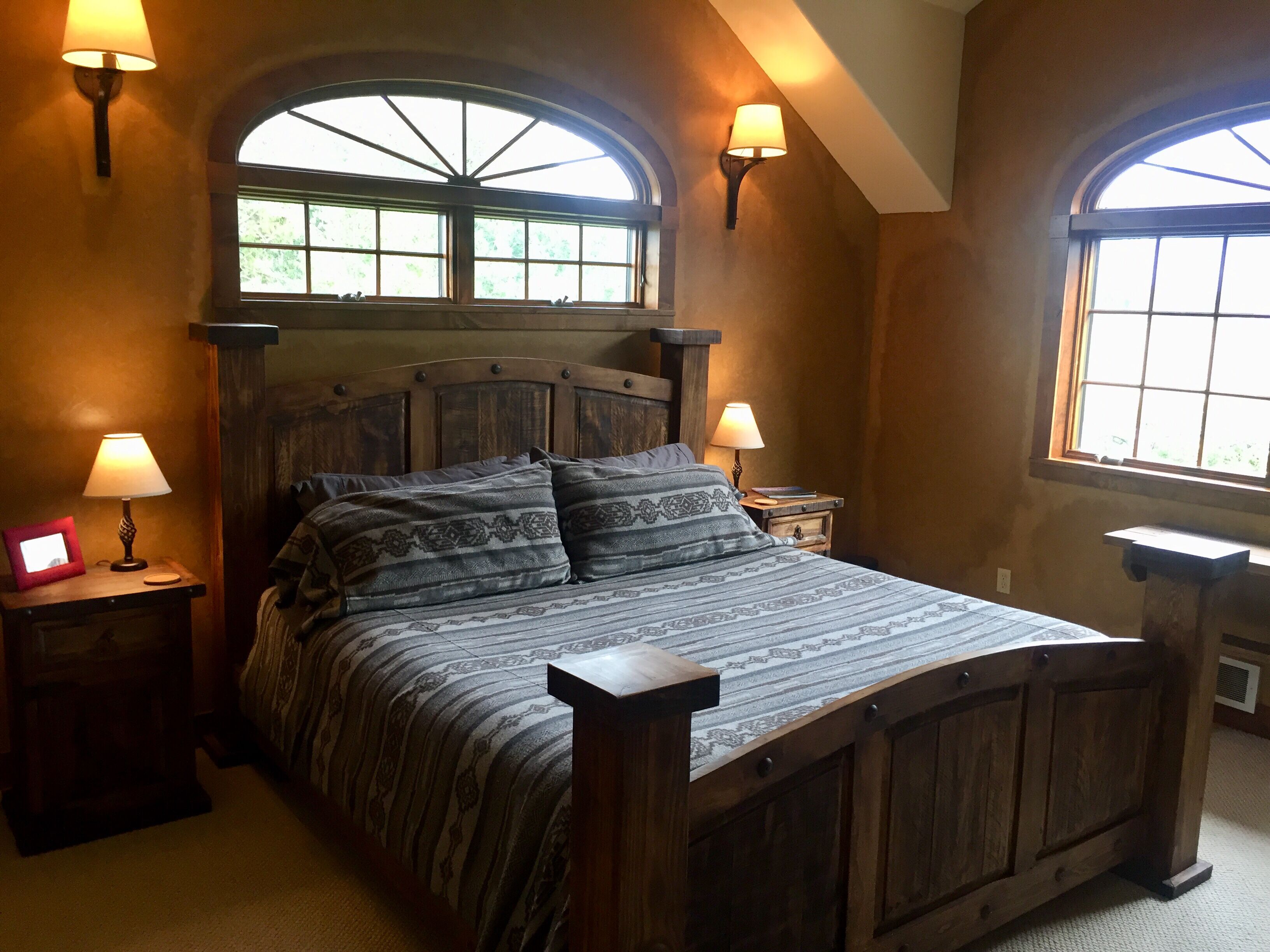 A bedroom with a wooden bed, two bedside lamps, and a large window with a view of greenery.