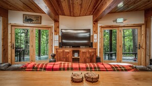 Ein gemütliches Blockhaus-Interieur mit einem großen Flachbildfernseher über einem Holztisch, an dessen Seite zwei Türen zu einer Waldterrasse führen.