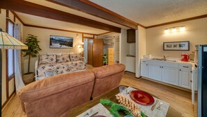 A cozy living room with a brown leather sofa, a dining table set with plates and cutlery, and a kitchen area with white cabinets and a refrigerator.