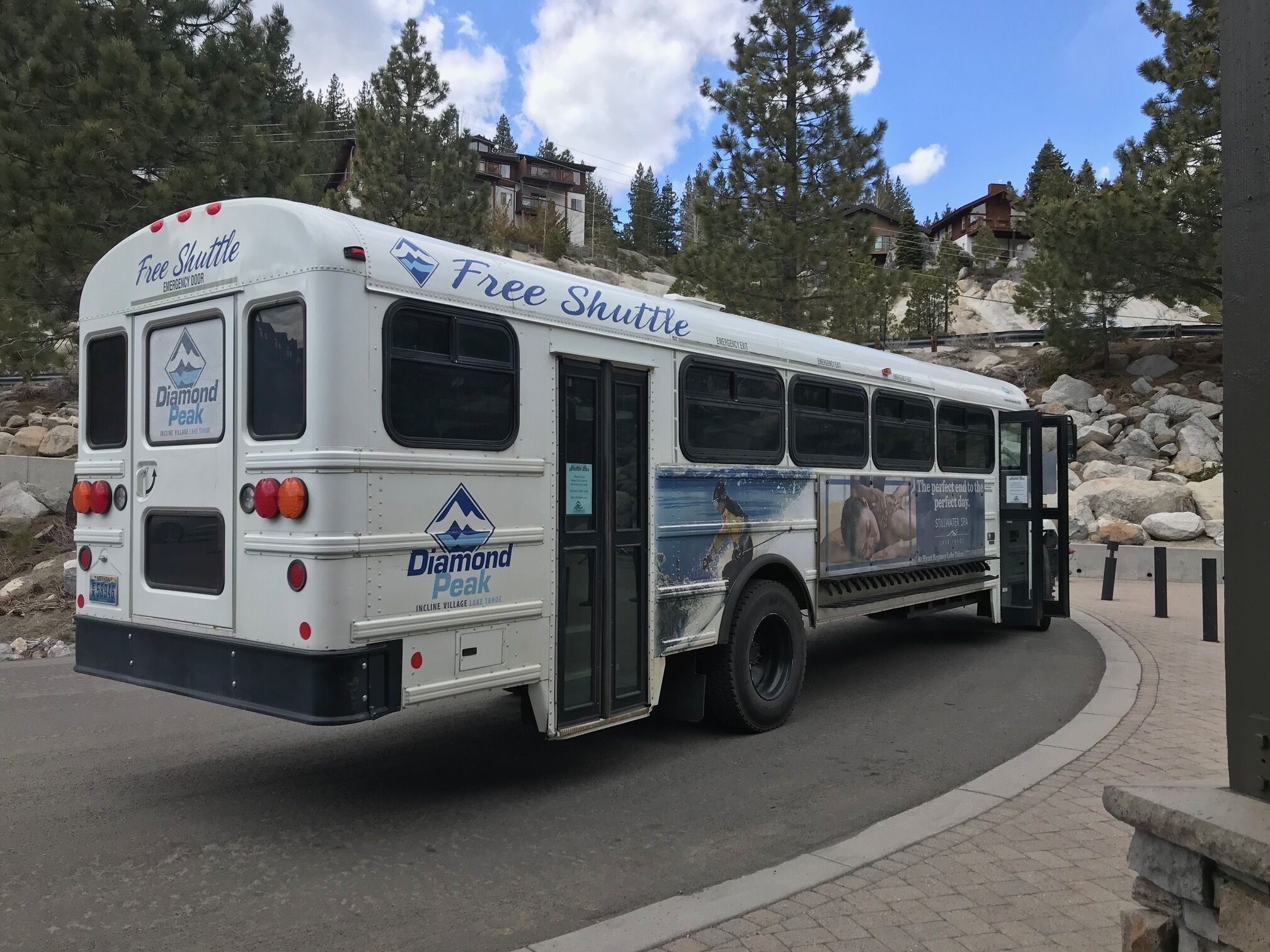 Un autobús lanzadera blanco con la leyenda "Free Shuttle" y el logotipo de "Diamond Peak", estacionado en una carretera con montañas al fondo.