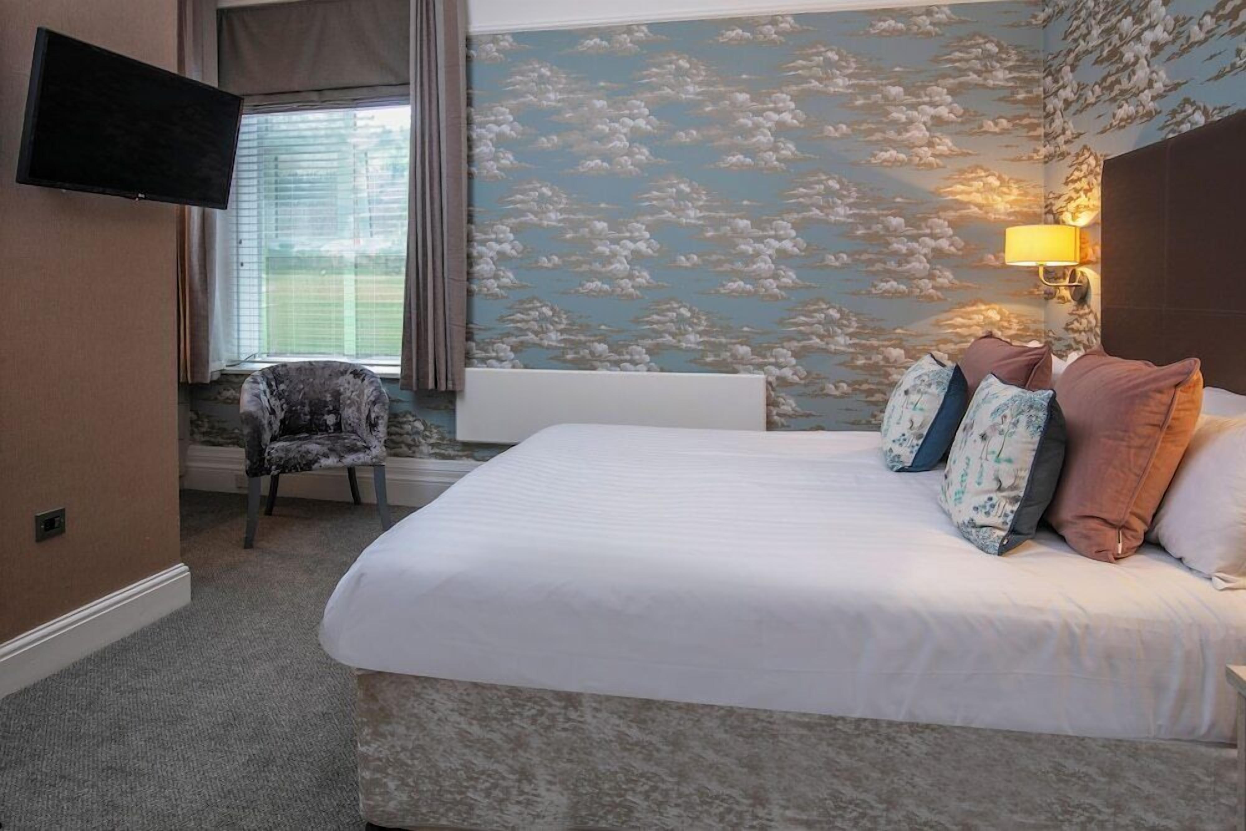 Habitación de hotel con cama, una silla, televisor y pared con diseño de nubes.