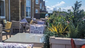 An outdoor seating area with wooden furniture and patterned cushions, surrounded by lush greenery and flowering plants.