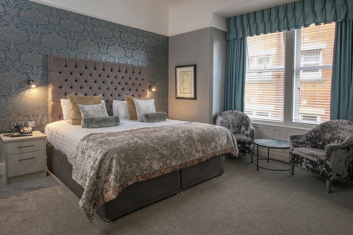 A bedroom with a tufted headboard, a large bed with multiple pillows, a nightstand, a chair, and a table.