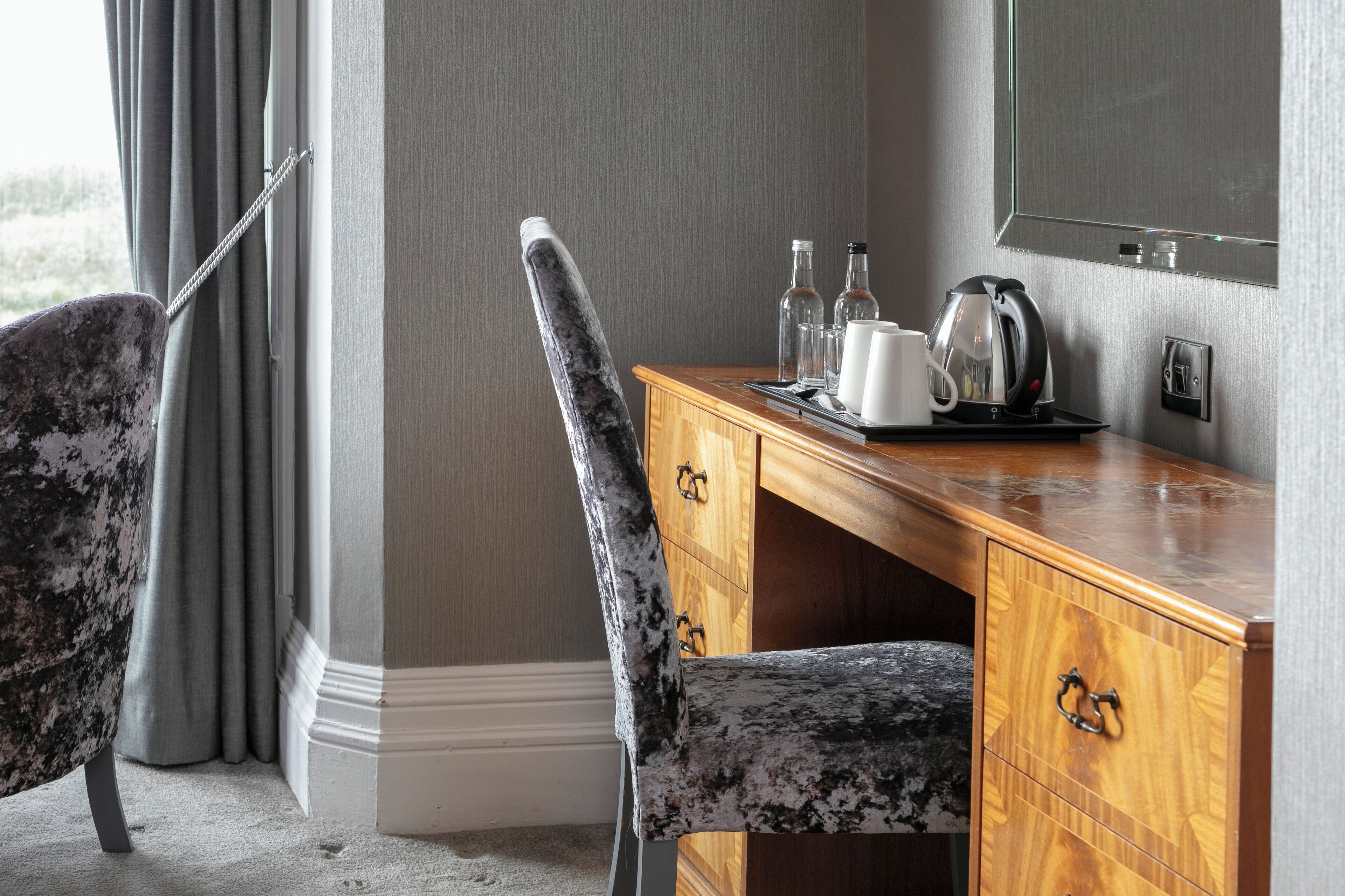 A hotel room with a wooden desk, a chair, a mirror, and a window with curtains.