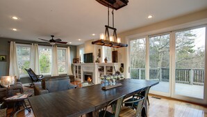 A spacious room with a large wooden dining table, comfortable seating, a fireplace, and large windows offering a view of the outdoors. - Burnt Ridge Retreat | Luxury Black Mountain Home w/ Game Room & Mountain Views (Black Mountain)