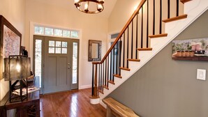 A staircase with wooden steps and black balusters, a gray door with a mirror above it, and a chandelier with multiple lights. - Burnt Ridge Retreat | Luxury Black Mountain Home w/ Game Room & Mountain Views (Black Mountain)