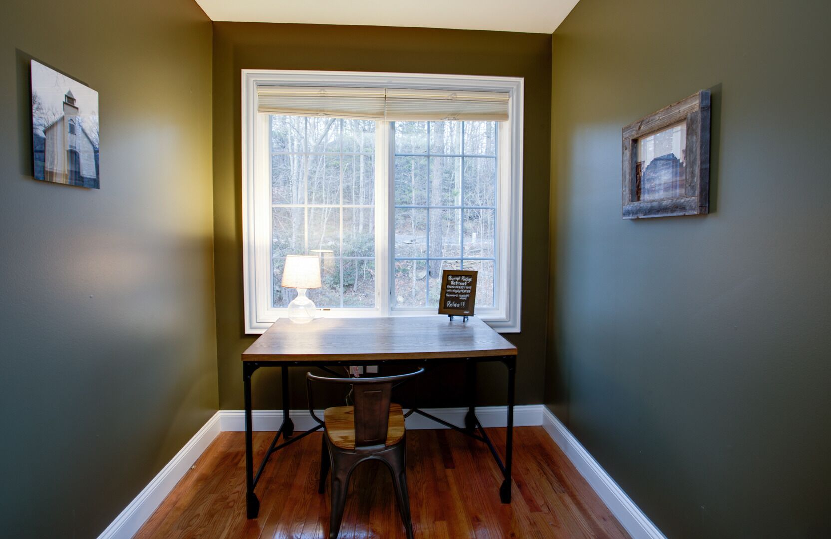 A room with a large window, a table with a lamp, and two framed pictures on the walls.
