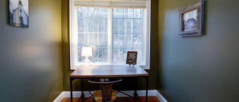 A room with a large window, a table with a lamp, and two framed pictures on the walls.