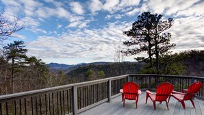 A deck with a railing, three red Adirondack chairs, and a view of a forested mountain range. - Burnt Ridge Retreat | Luxury Black Mountain Home w/ Game Room & Mountain Views (Black Mountain)