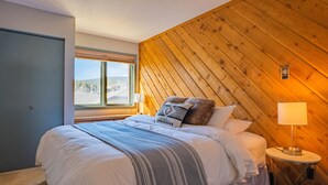 Un dormitorio con cama, ventana, pared de madera y mesita de noche con lámpara.