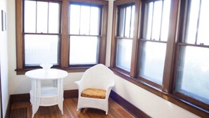 A room with a white wicker chair and a small round table, large windows with wooden frames, and a wooden floor.