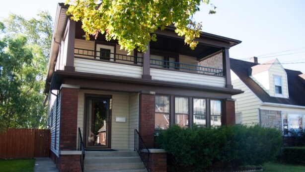 A two-story house with a front porch, a balcony on the upper floor, and a brick foundation.