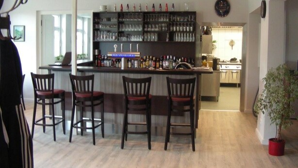 A bar area with a counter, multiple shelves of bottles, and four bar stools. - Hotel Langstedt (Langstedt)