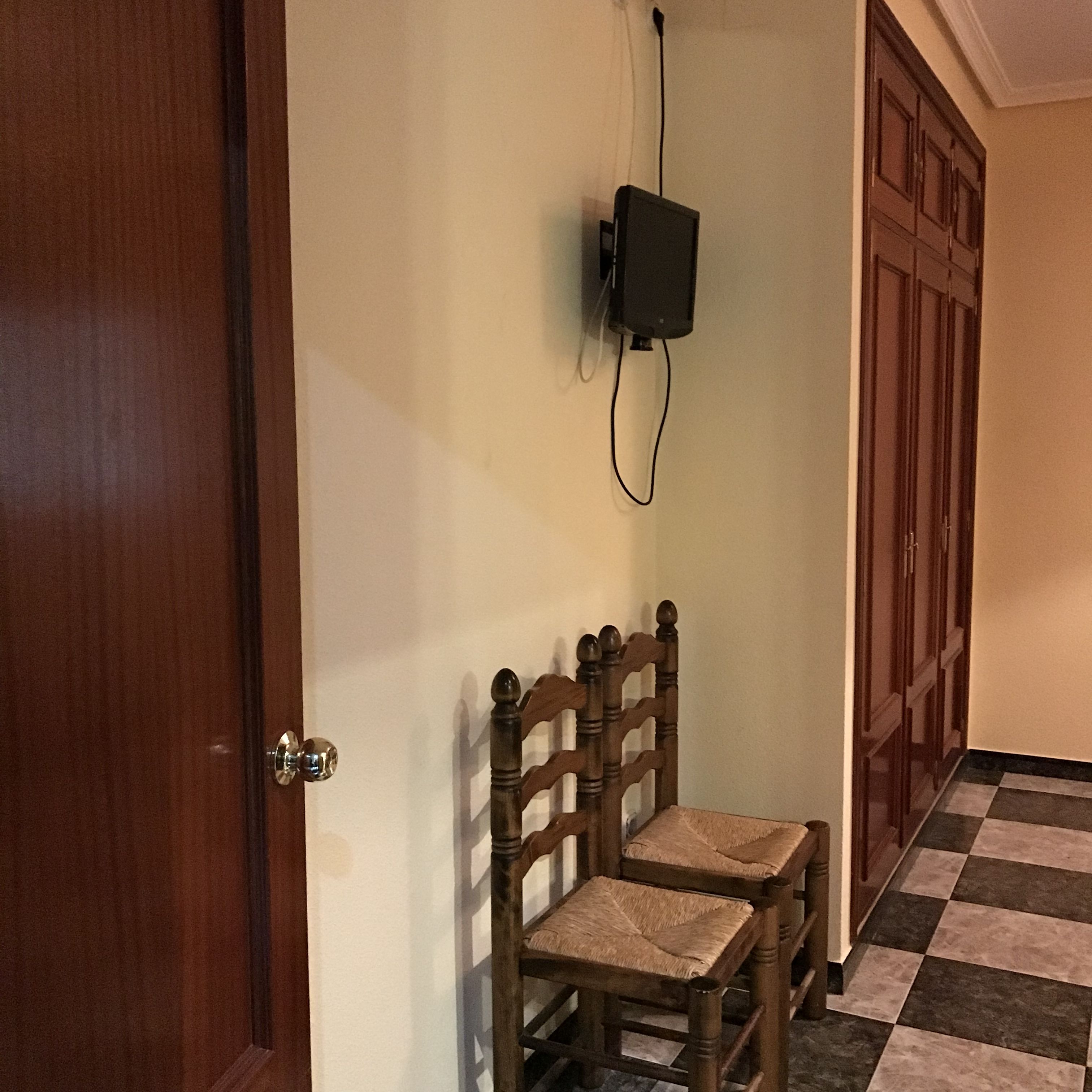 Un corridoio con una porta in legno, una sedia di legno con seduta intrecciata e un altoparlante nero fissato al muro.