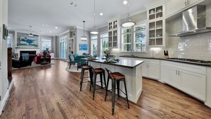 A spacious kitchen with white cabinets, a large island with stools, pendant lighting, and a view of the living area.
