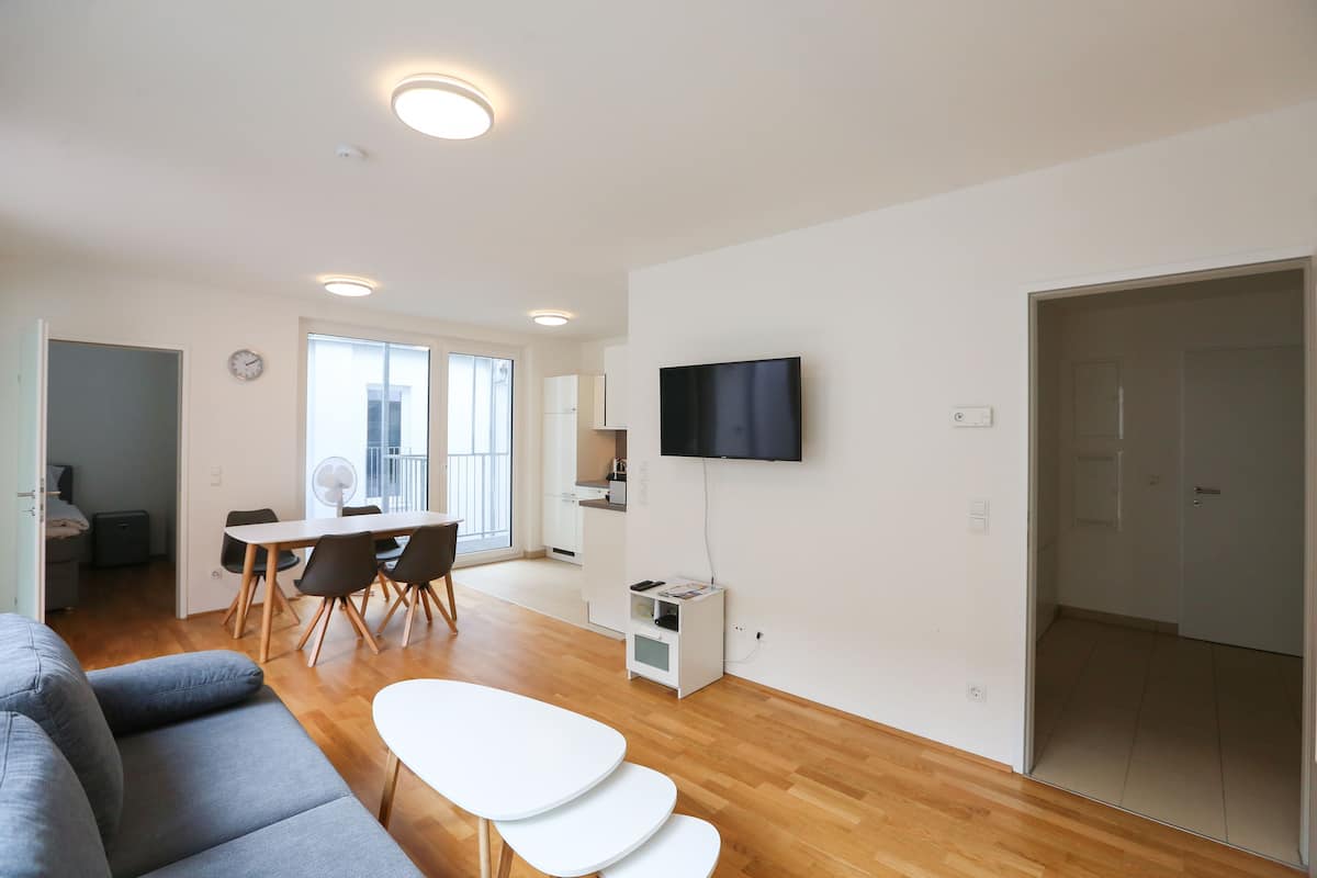 A modern living room with a grey sofa, a white dining table, and a flat-screen TV mounted on the wall.