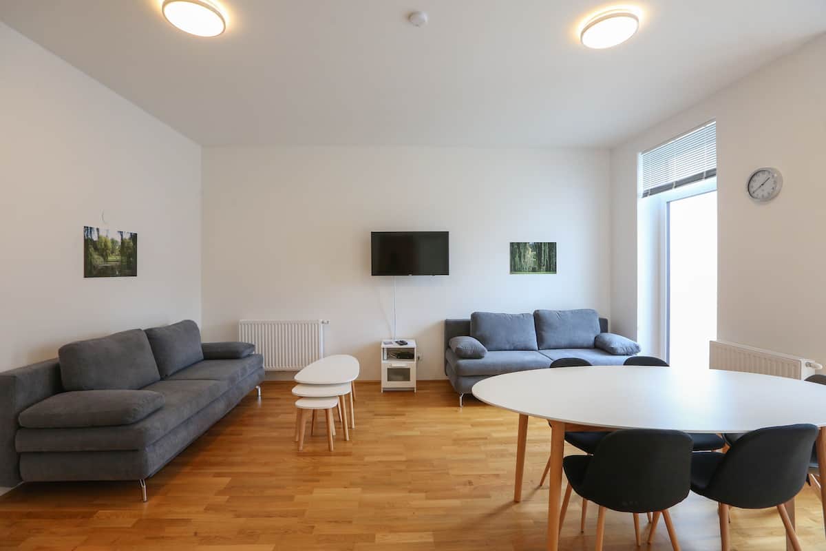 A modern living room with a grey sofa, a white dining table, and a flat-screen TV mounted on the wall.