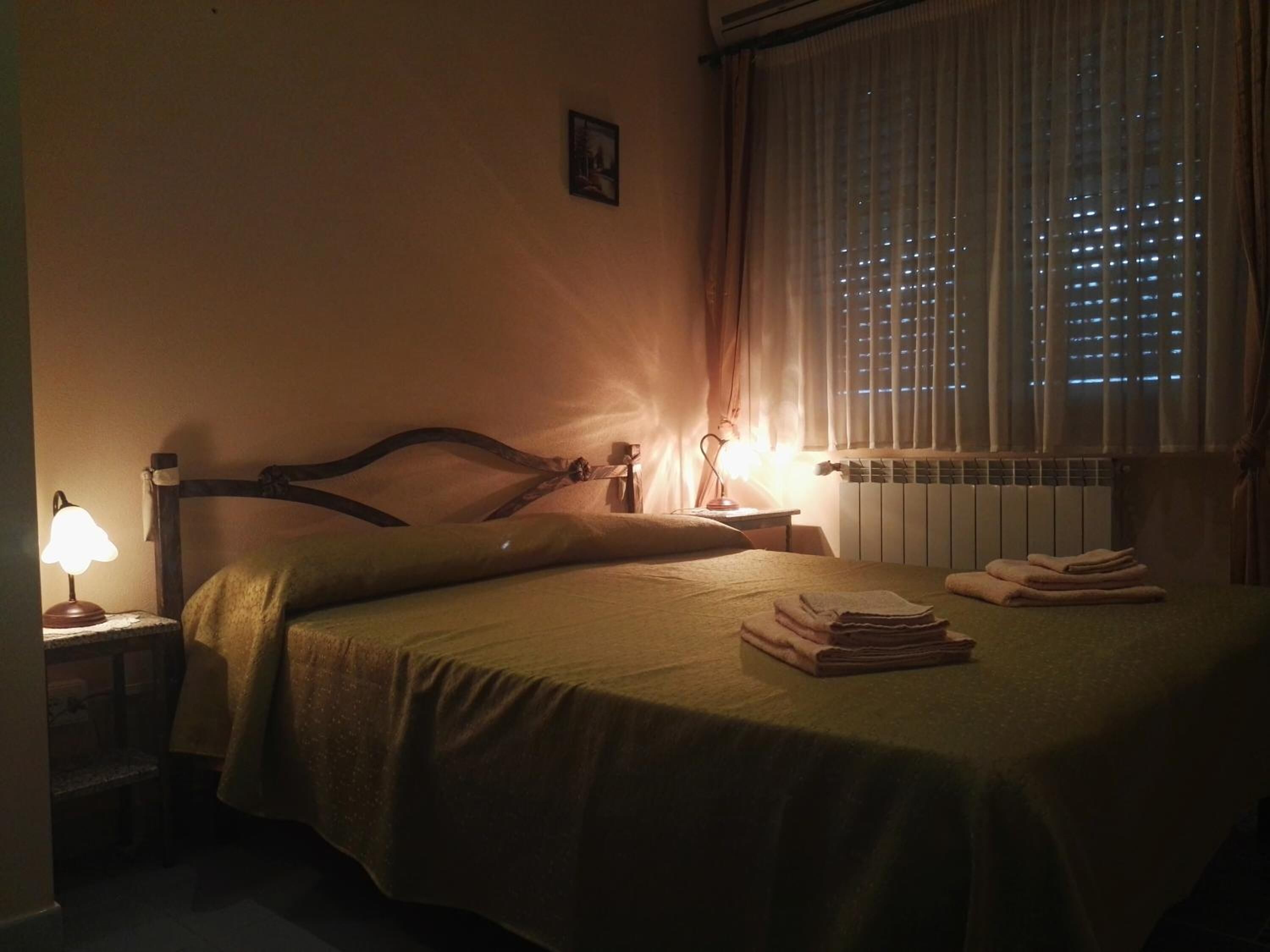 Ein schummrig beleuchtetes Hotelzimmer mit Bett, Nachttisch, Lampe und einem Fenster mit Vorhängen.