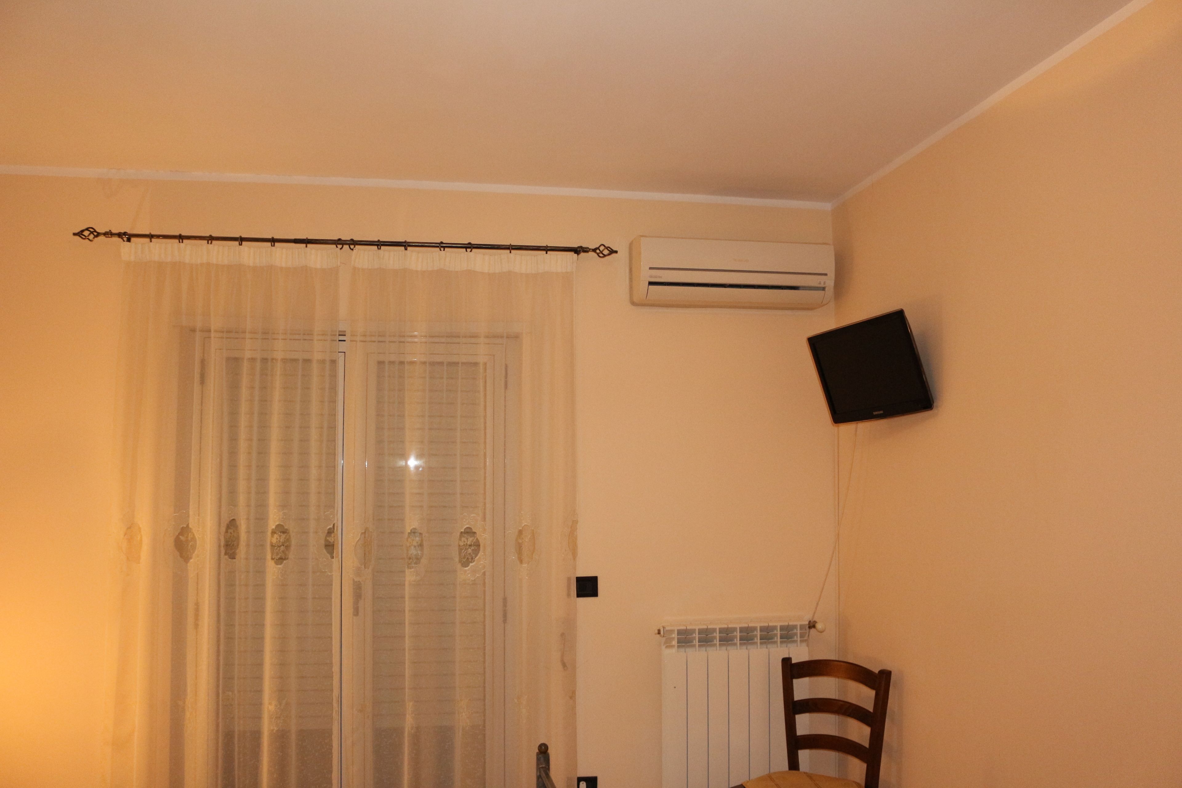A room with a window covered by sheer curtains, an air conditioning unit mounted on the wall, and a television mounted on the wall.