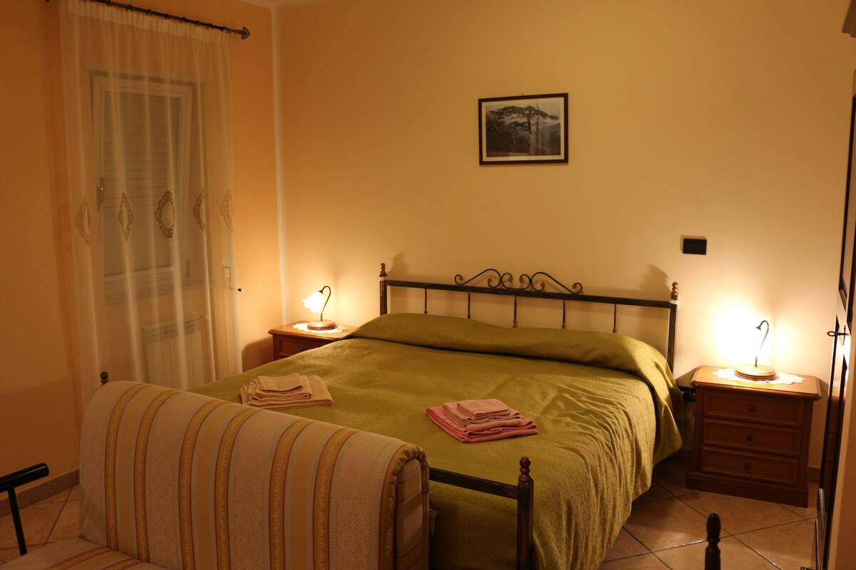 A bedroom with a bed, two bedside tables, a nightstand, a chair, a window with curtains, and a framed picture on the wall.
