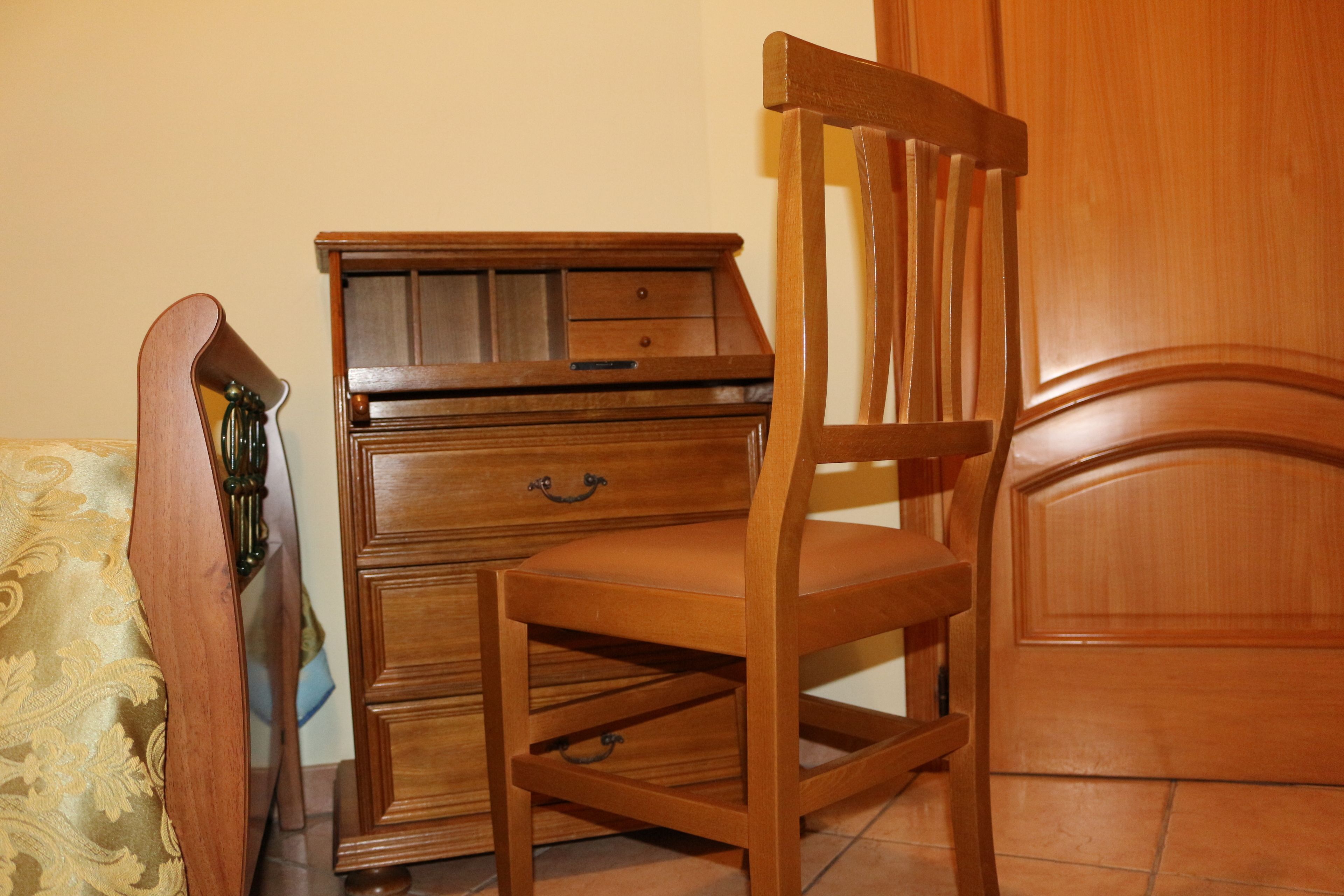 Ein Holzhocker mit gepolstertem Sitz und Rückenlehne, platziert neben einer Holzkommode mit Schubladen und einer kleinen Ablagefläche.