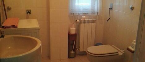 Une petite salle de bain avec un lavabo, des toilettes et un bidet. On y trouve une fenêtre avec des stores, un porte-serviettes et un radiateur.