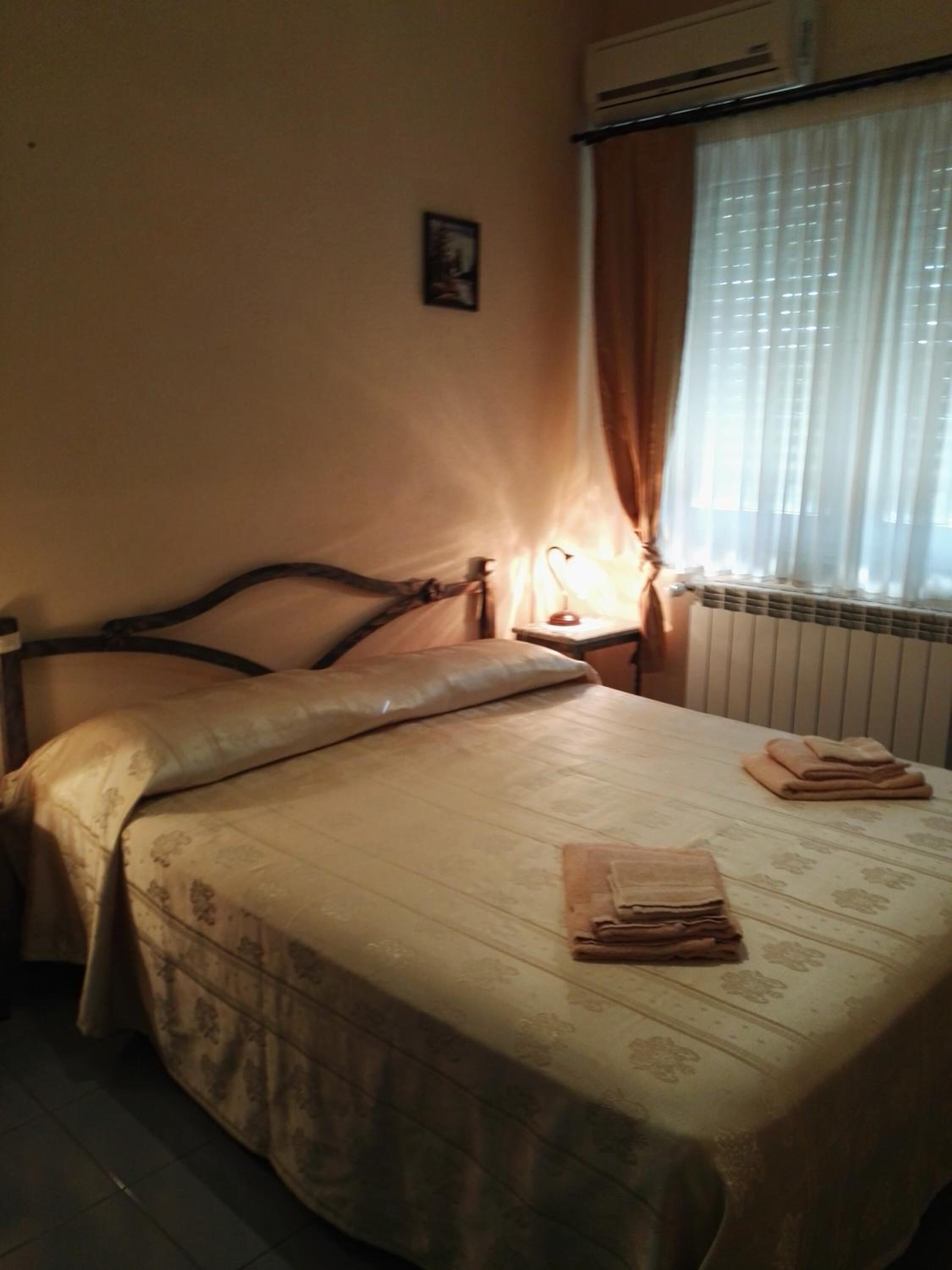 A bedroom with a bed, a nightstand, a lamp, a radiator, and a window with curtains.
